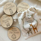 A set of milestone wooden discs for babies, each featuring animal illustrations and inscriptions for "One Month," "Two Months," "Three Months," and "Hello World, I'm Jacob." The discs are accompanied by a fabric pouch decorated with animal illustrations and the words "Jacob’s First Year. Memories & moments."