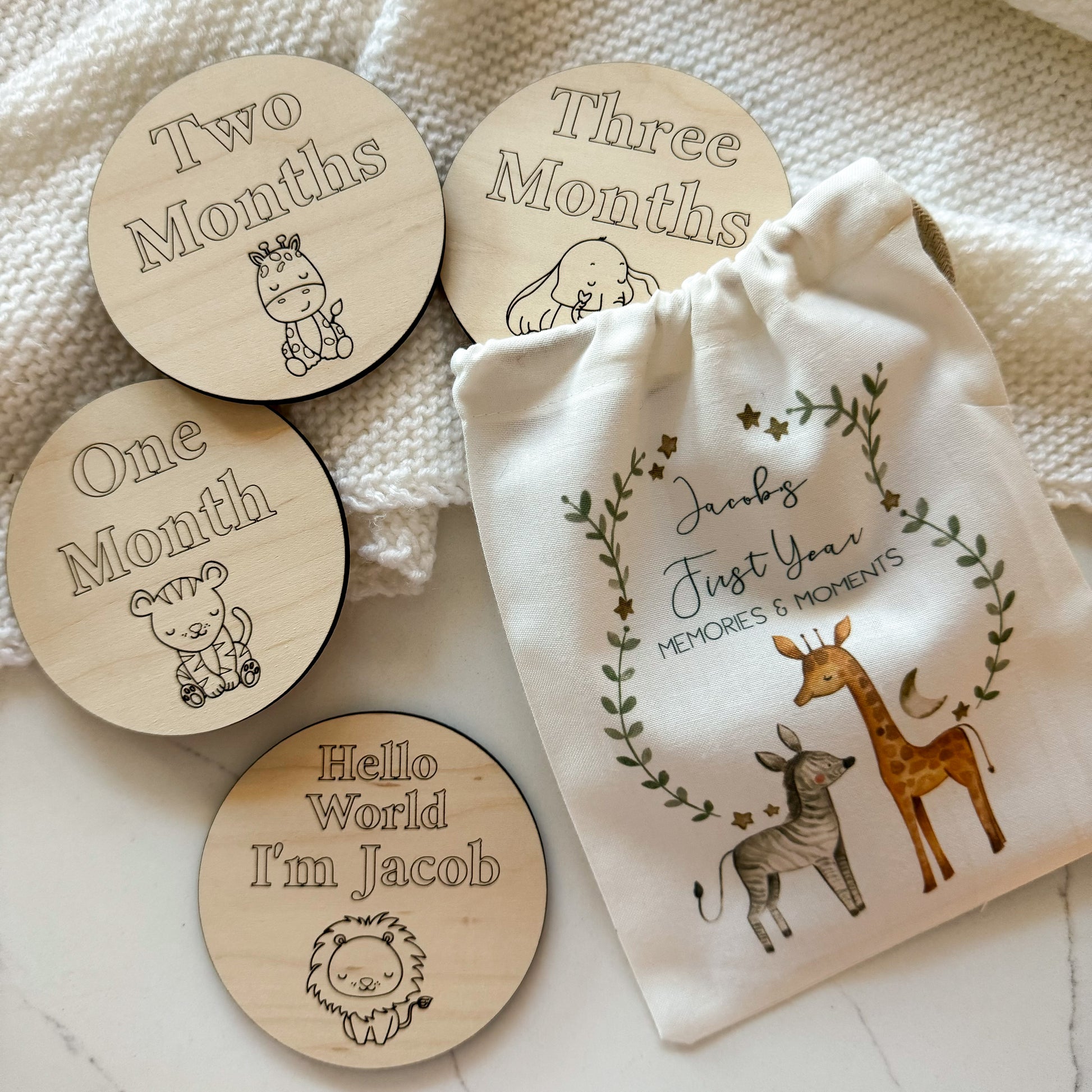 A set of milestone wooden discs for babies, each featuring animal illustrations and inscriptions for "One Month," "Two Months," "Three Months," and "Hello World, I'm Jacob." The discs are accompanied by a fabric pouch decorated with animal illustrations and the words "Jacob’s First Year. Memories & moments."